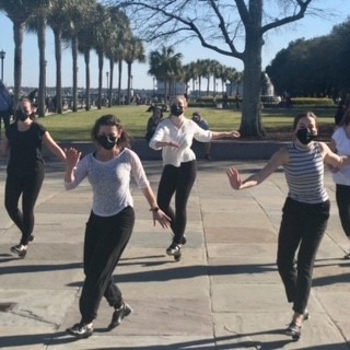 Tap dancers in white tops and black pants perform in a beautiful park lined with palmetto trees.